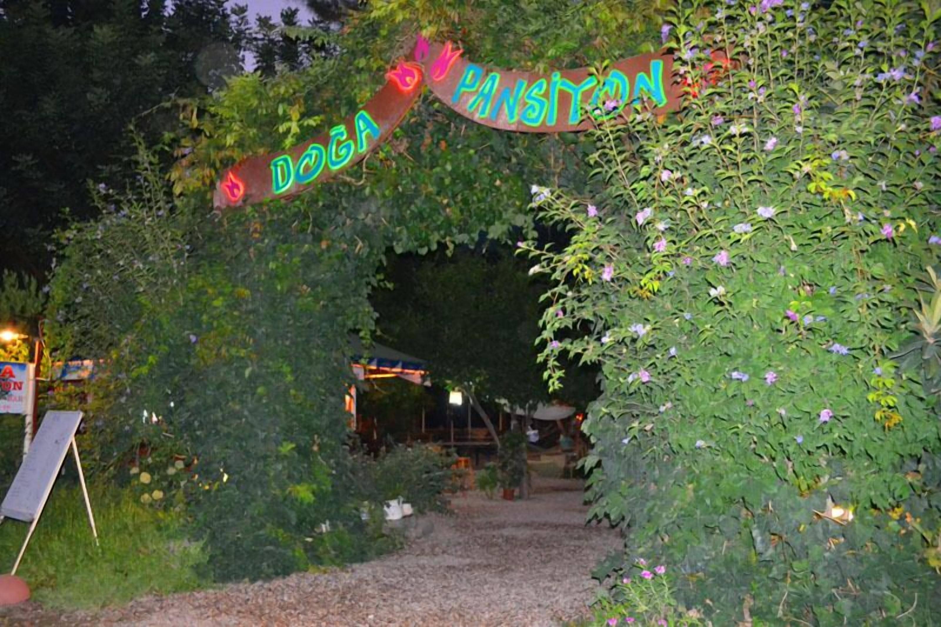 Une entrée ornée d’un panneau néon sur lequel on peut lire « DOGA PANSYON », le tout au milieu d’une végétation luxuriante.