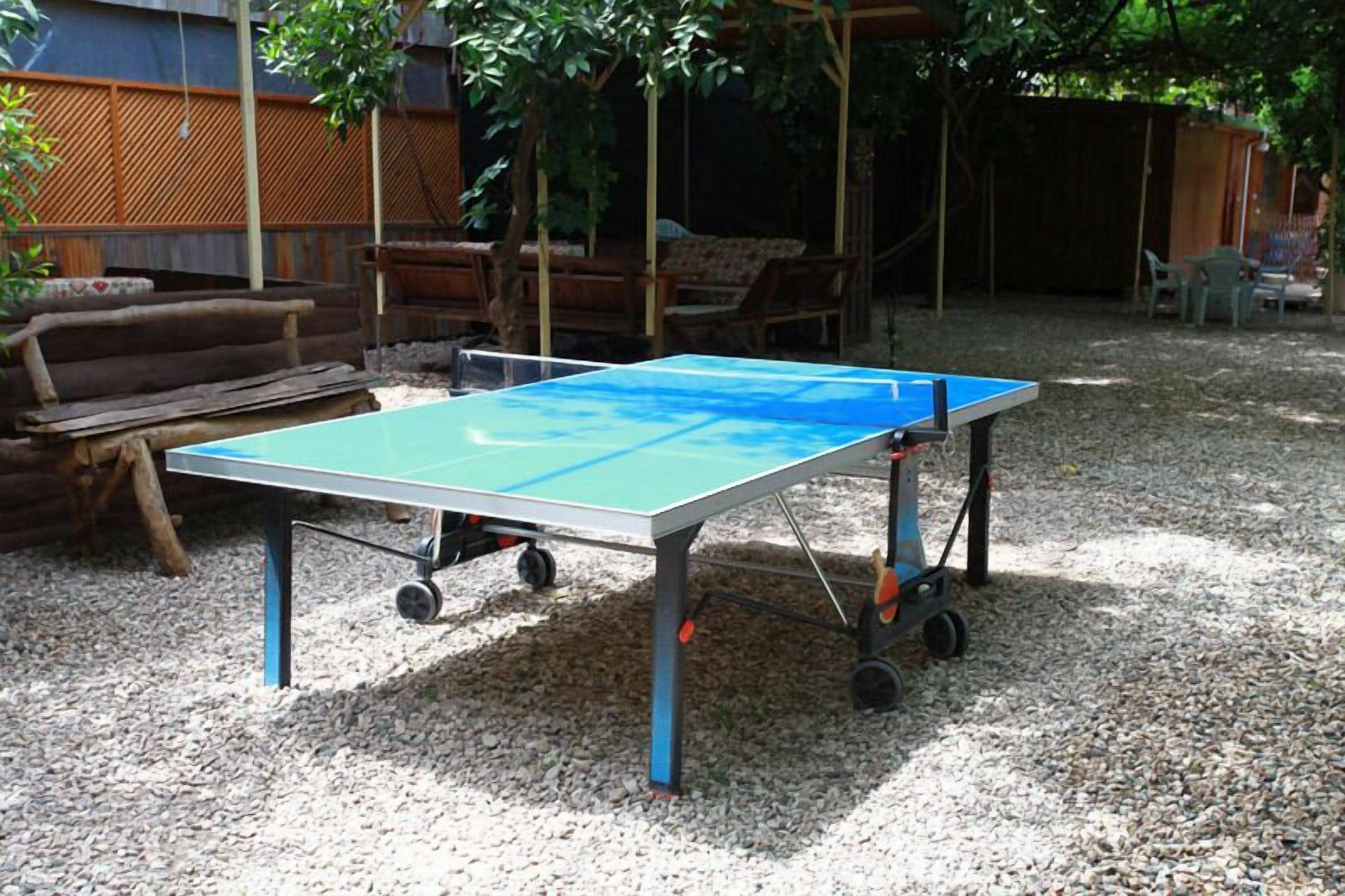 A ping pong table in an outdoor area with gravel ground, wooden furniture, and a wooden fence in the background.