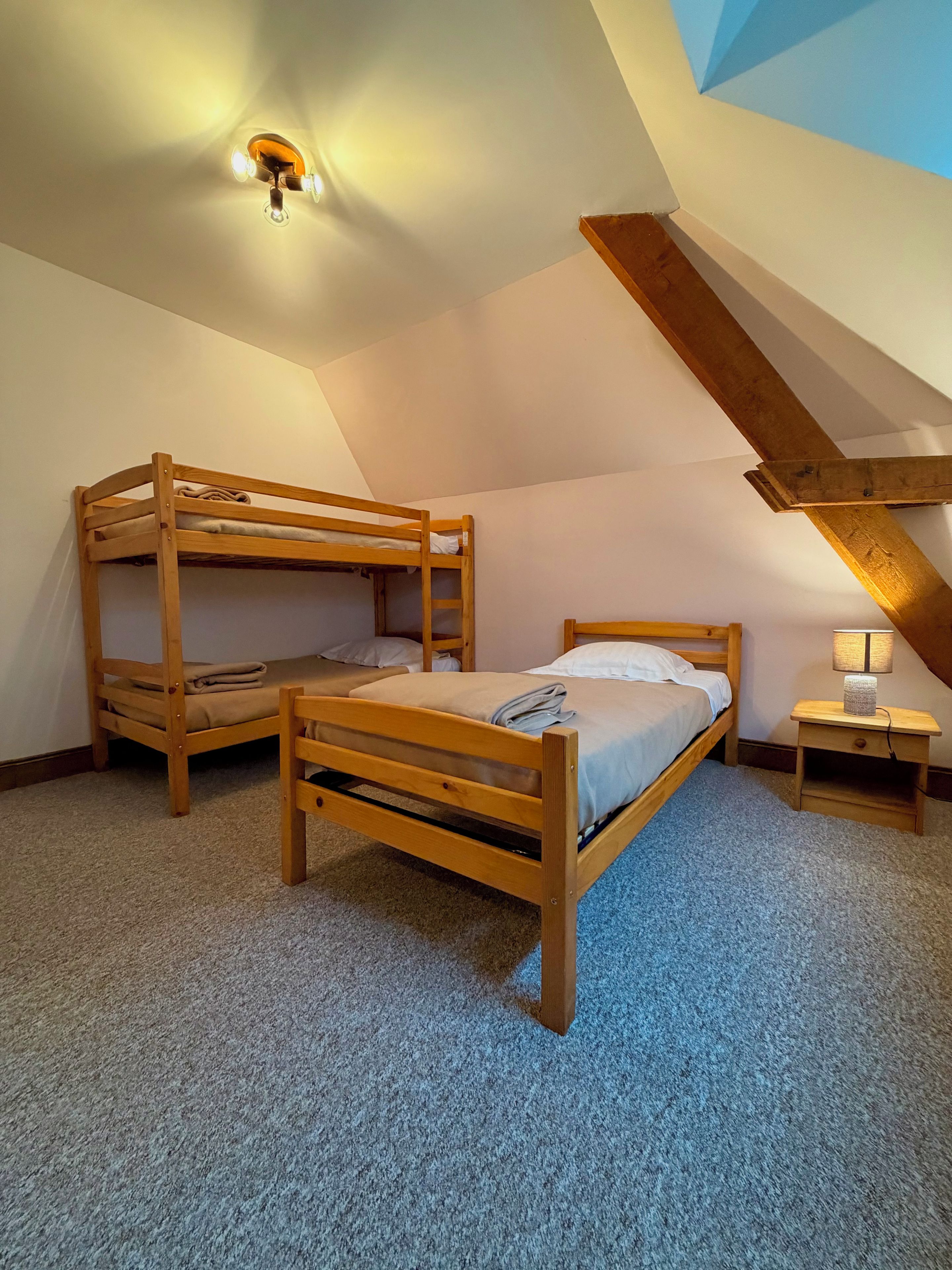 Una habitación con dos camas literas de madera, una cama individual, una mesita de noche con lámpara y un techo inclinado con luminaria.