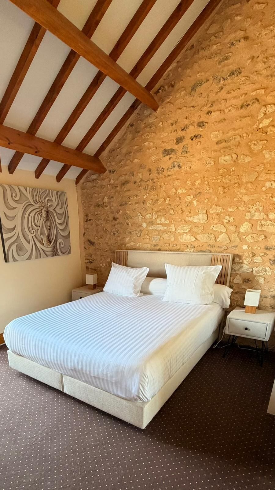 Un dormitorio con pared de piedra, cama con ropa de cama blanca y mesita de noche con lámpara.