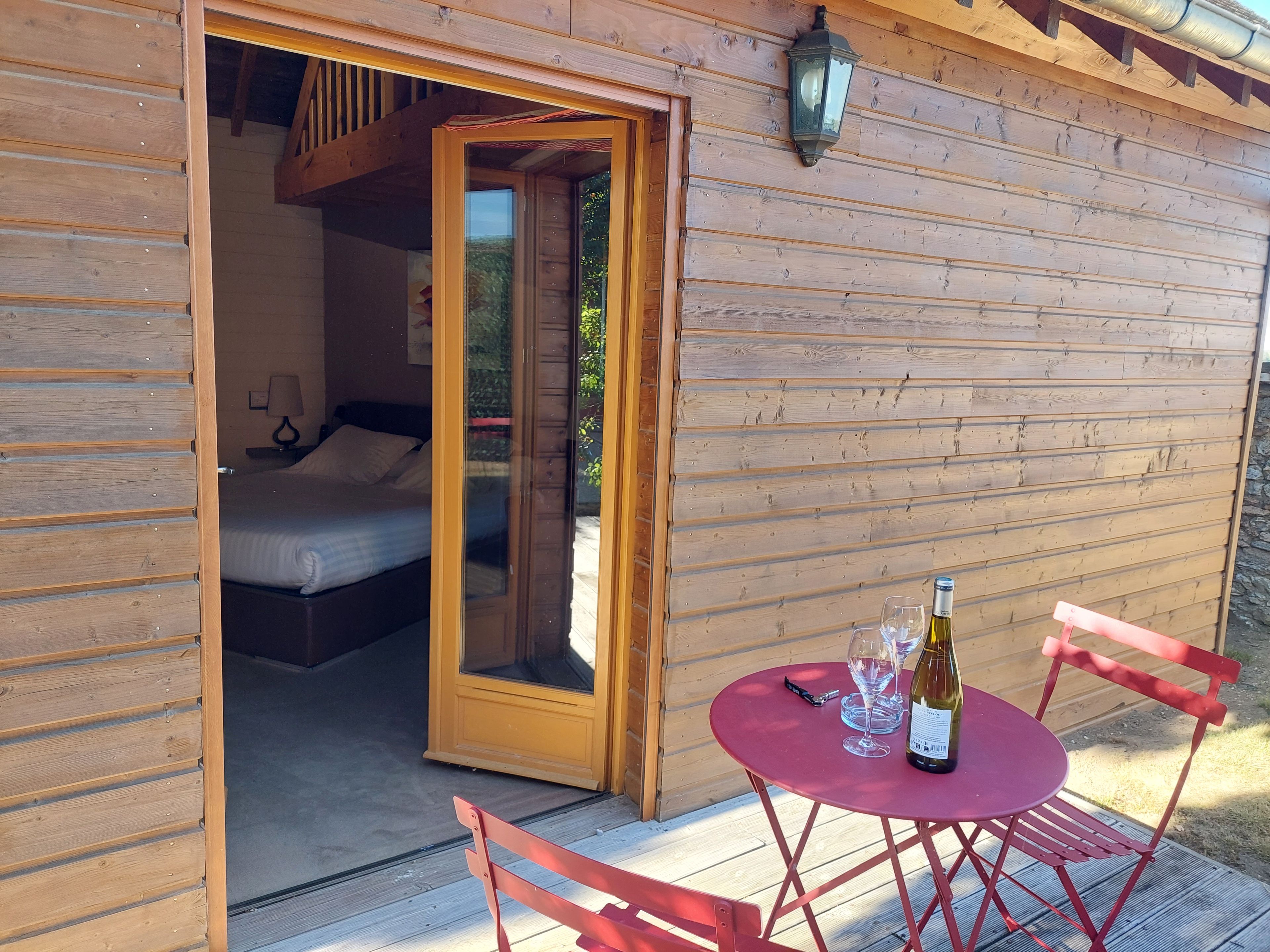 A wooden cabin with a bed, a table with wine glasses and a bottle, and a red chair outside.