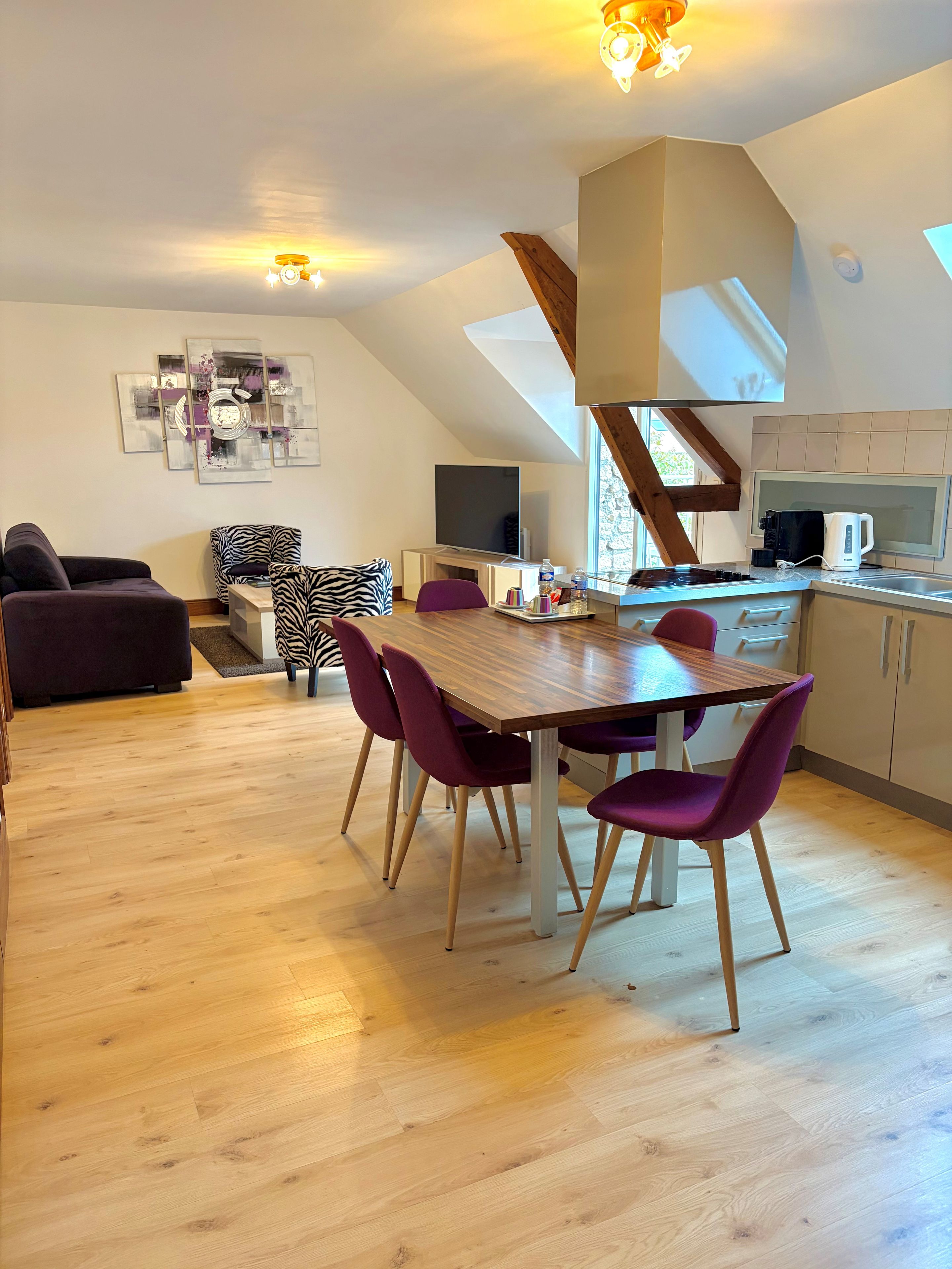 A modern kitchen with a wooden table and purple chairs, a dining area with a sofa and zebra-patterned chair, and a TV on a white cabinet.