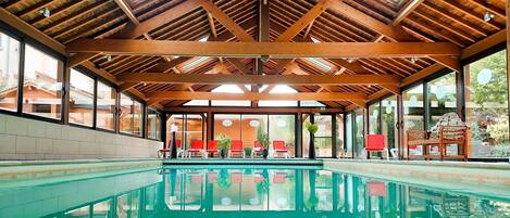 Piscina techada con vigas de madera y paredes de vidrio, rodeada de sillas rojas y plantas en macetas.