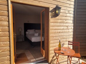 Una cabaña de madera con un dormitorio que incluye una cama, una mesita de noche y una lámpara. En el exterior, hay una mesa con una botella y vasos, y una silla.