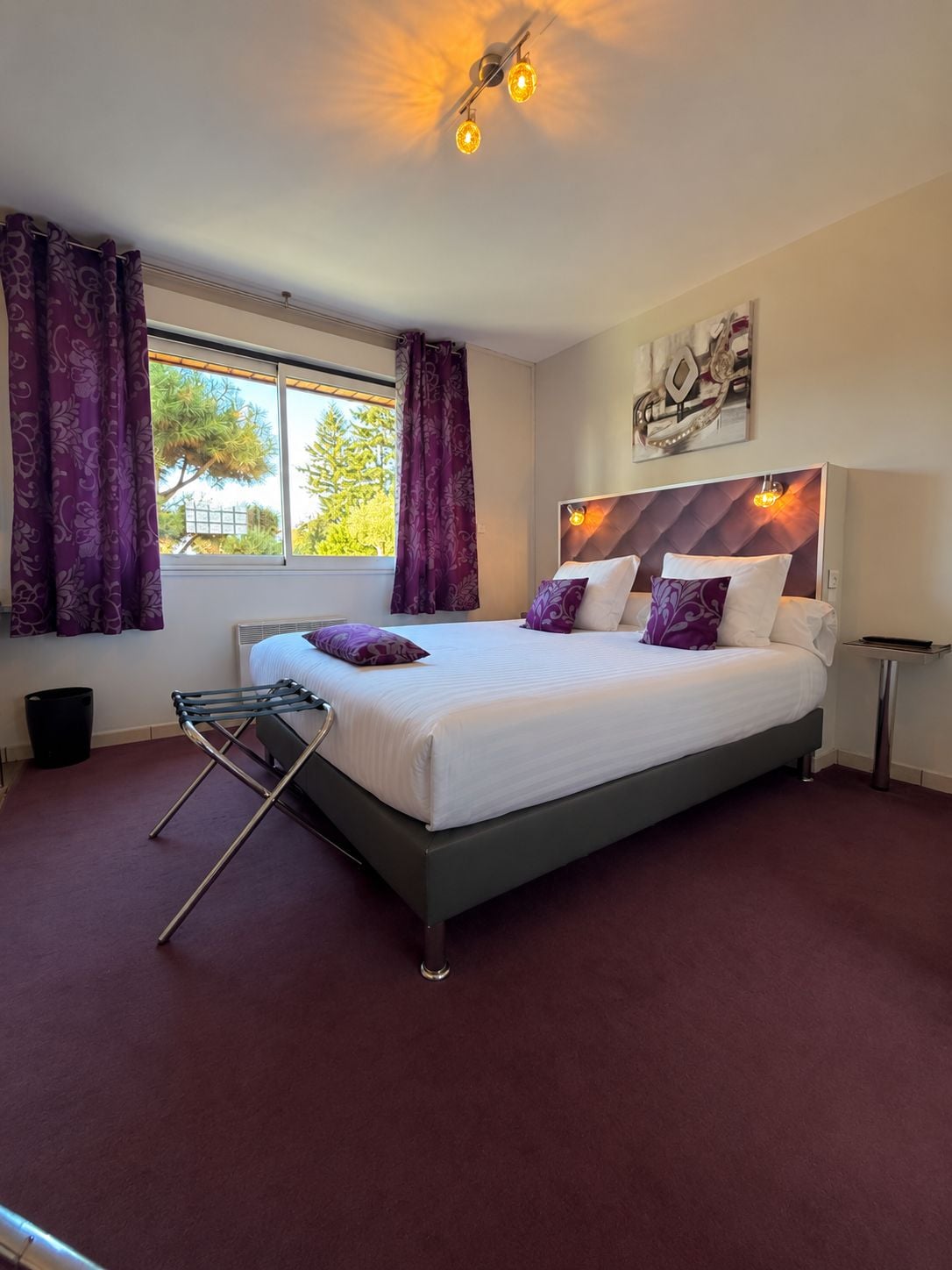 A bedroom with a bed, a window with curtains, a radiator, and a wall-mounted light fixture.