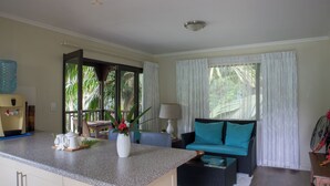 Una sala de estar moderna con zona de cocina, ventanas amplias con cortinas, un sofá con cojines color turquesa y una mesa de centro.