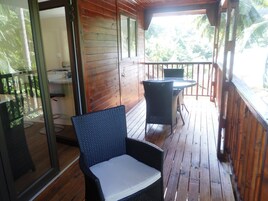 Una terraza de madera con mesa y sillas, una puerta de vidrio que da al interior, y vistas de follaje verde al exterior.