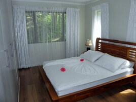 Una cama bien hecha con sábanas blancas y cabecera de madera, una mesita de noche con lámpara y una ventana con cortinas translúcidas.