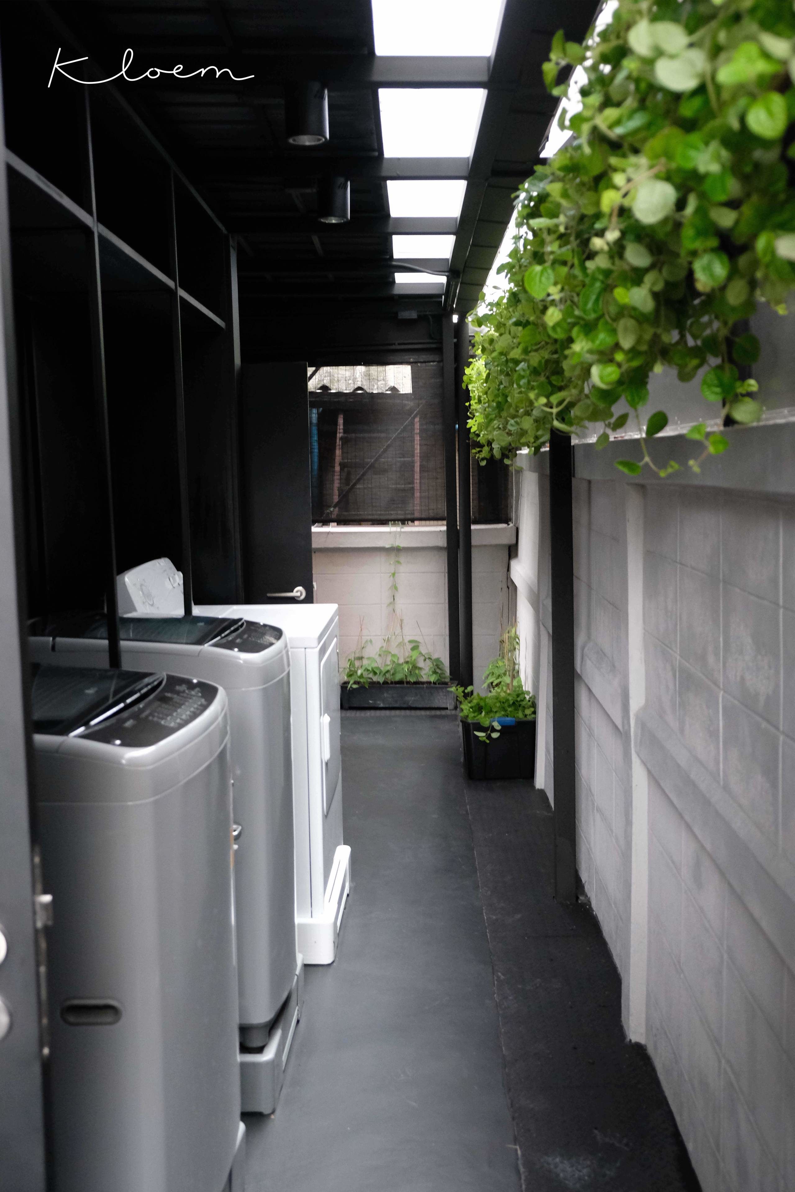 A narrow corridor with two stacked washing machines, a small garden area with potted plants, and a view of a building in the distance.