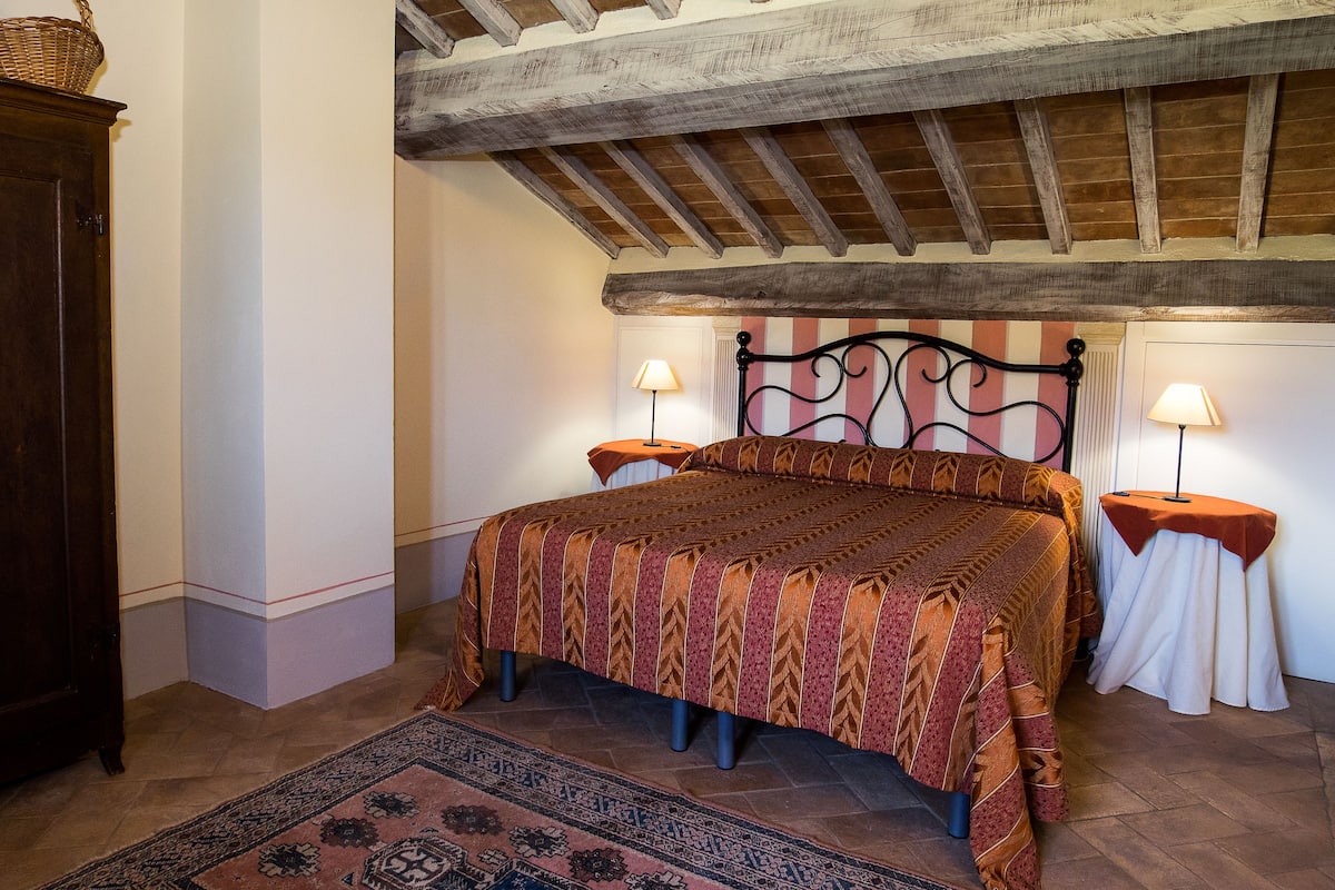 Un dormitorio con techo de madera, una cama grande con colcha estampada, dos mesitas de noche con lámparas y un armario de madera oscura.