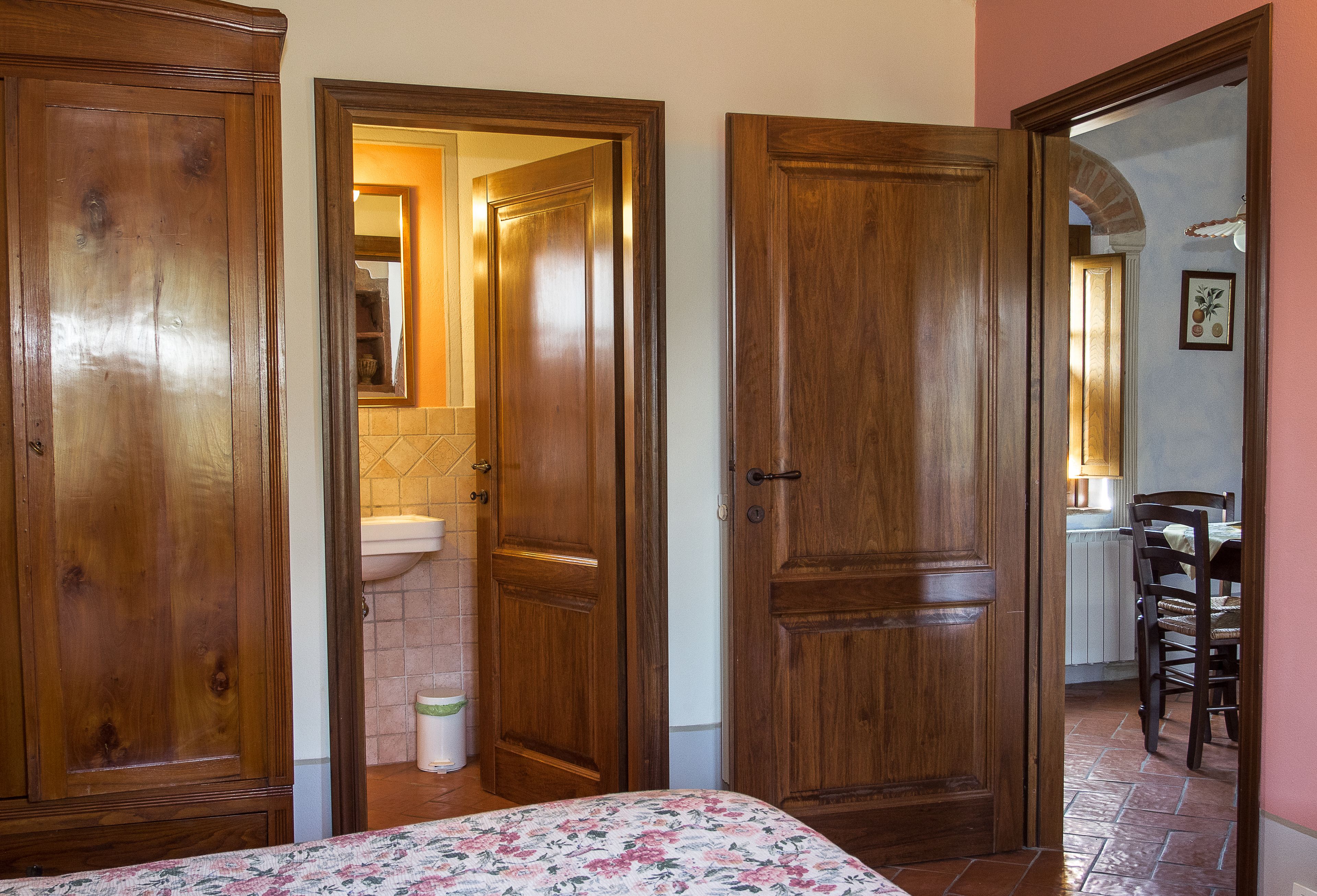Una camera da letto con porte in legno, un letto con copripiumino floreale, un bagno con lavandino e specchio, e una zona pranzo con sedie e tavolo.