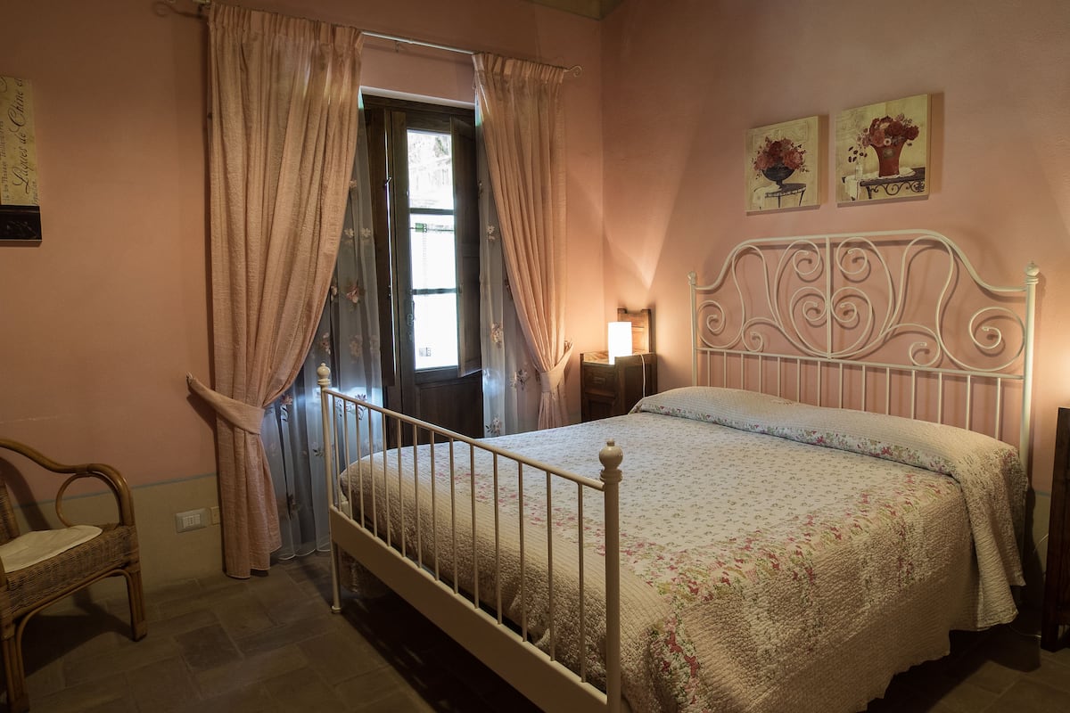 Un dormitorio con cama, una silla, una mesita de noche y una ventana con cortinas.