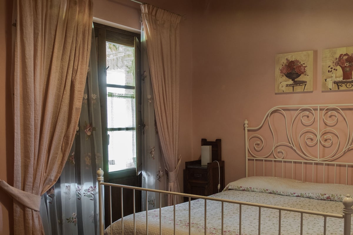 Un dormitorio con una cama, cortinas, una mesita de noche y dos cuadros colgados en la pared.