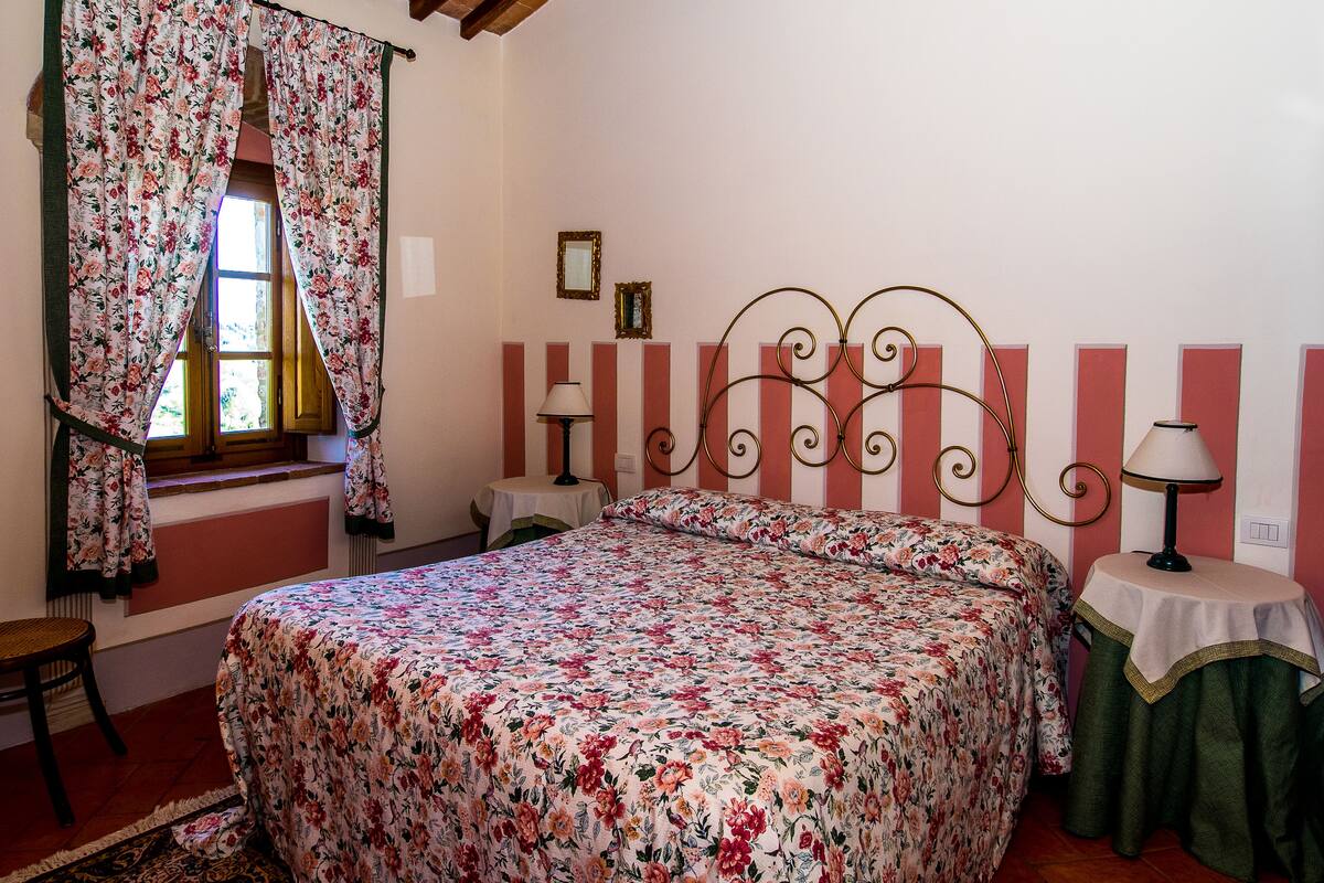 Un dormitorio con colcha estampada, una ventana con cortinas, una mesita de noche con lámpara y una silla.