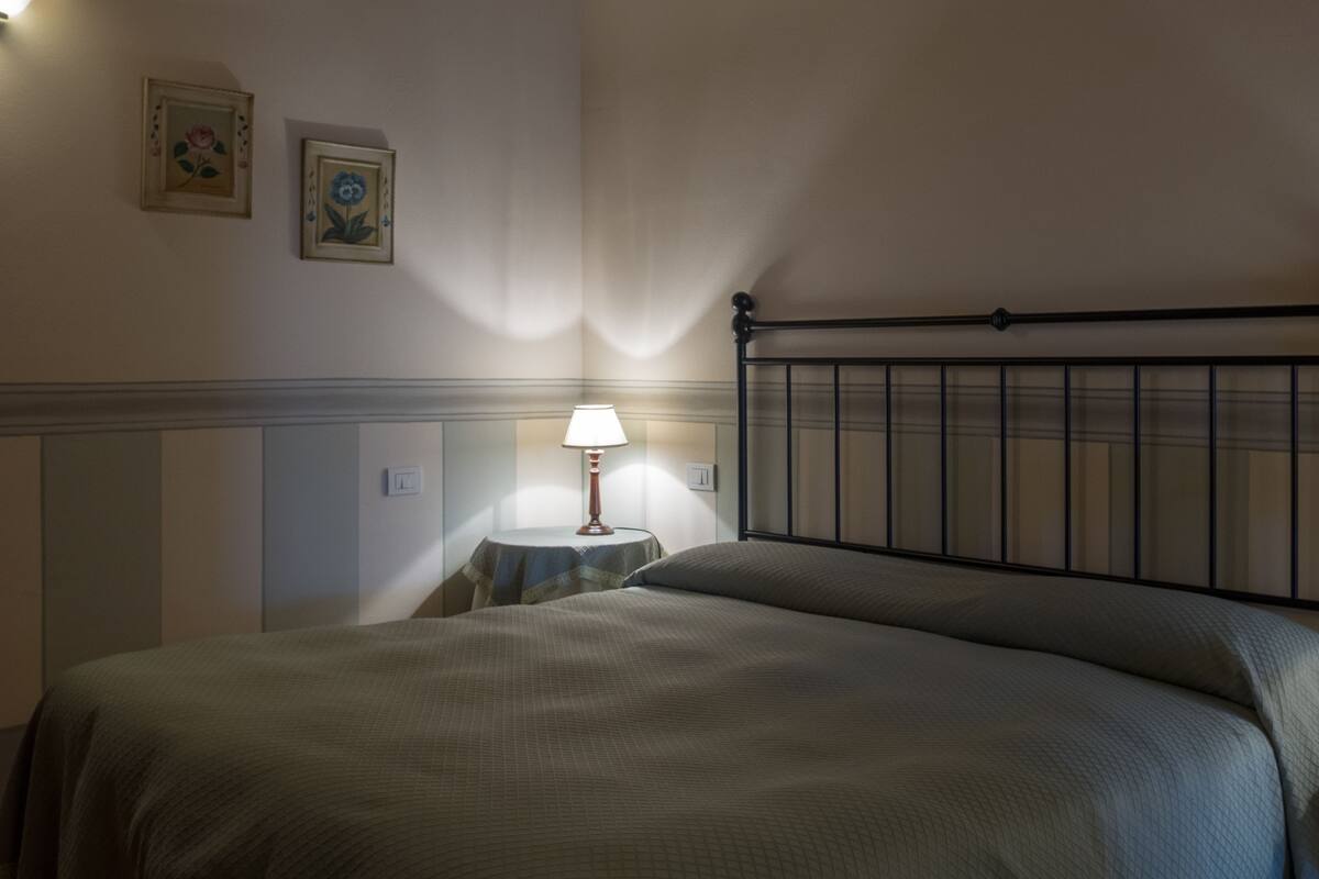 Una cama bien hecha con cabecera, una mesita de noche con lámpara y dos cuadros enmarcados en la pared.