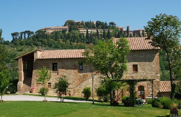 A stone building with a tiled roof, surrounded by a well-maintained garden and a view of a hilltop castle. - Agriturismo Nobile (Montepulciano)