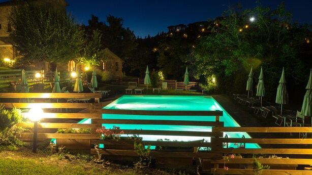 Une piscine rectangulaire entourée d’une terrasse en bois, illuminée par des lumières chaleureuses.
