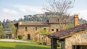 Un bâtiment en pierre avec un toit en tuiles, une cour pavée et une vue sur une colline parsemée d’arbres, avec un château visible au loin.