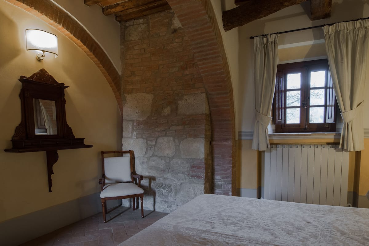 Un dormitorio con pared de piedra, una silla de madera, una cama, un ventanal con cortinas y una lámpara de pared.