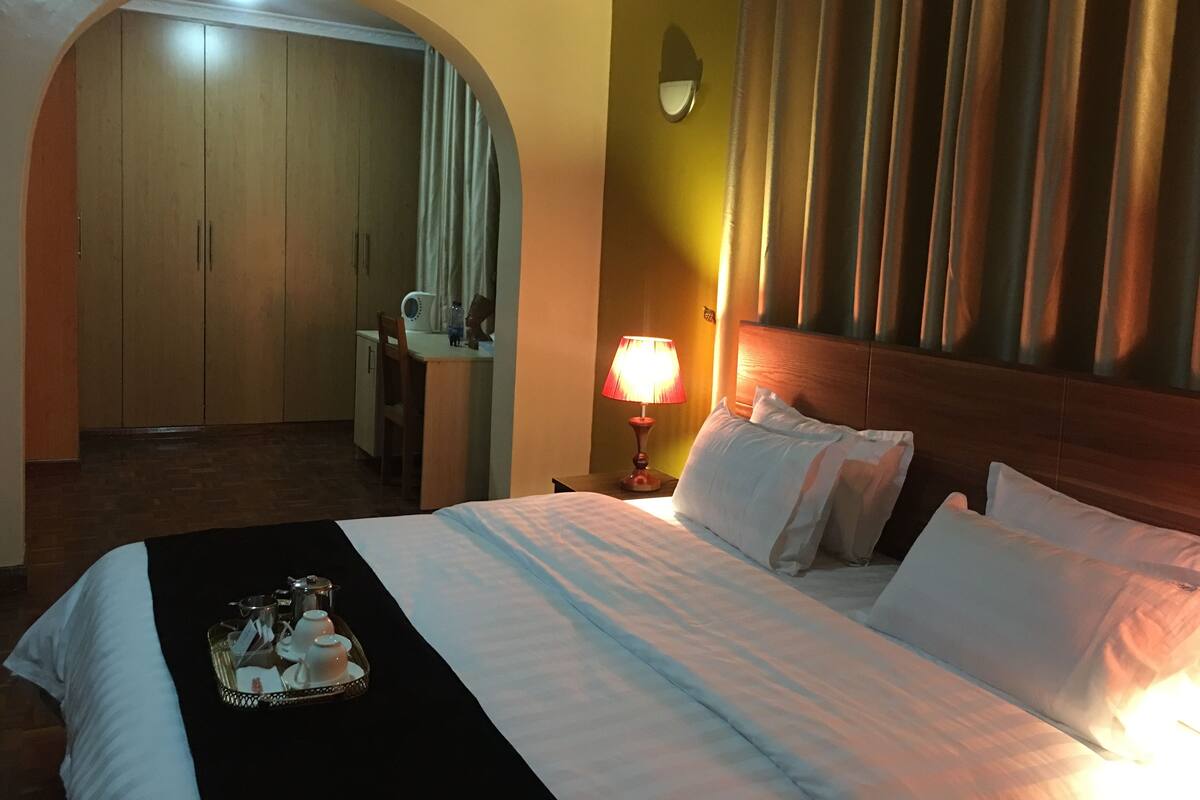 Habitación de hotel con cama, mesita de noche con lámpara, bandeja con tazas de té, espejo y armario.