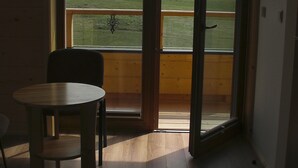 Una habitación con piso de madera, una mesa redonda y una silla. Se aprecia un ventanal con vistas a un campo cubierto de hierba y colinas.