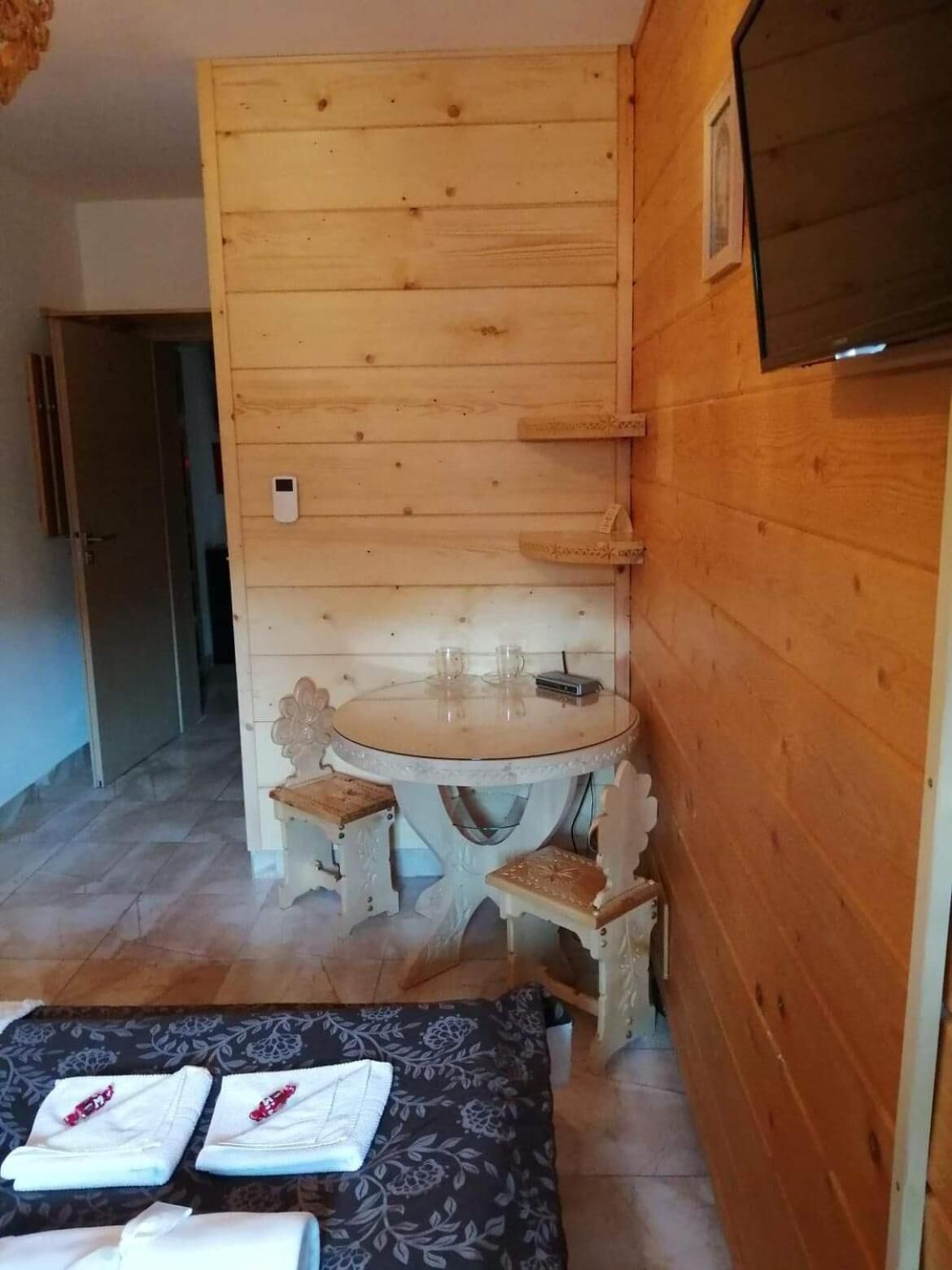 A wooden room with a round table and chairs, a bed with neatly folded towels, and a door leading to another room.