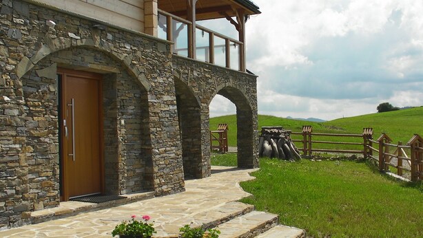 Un edificio de piedra con puerta de madera y ventanas arqueadas, un camino de piedra que lleva a la entrada, y un área con césped y valla de madera.