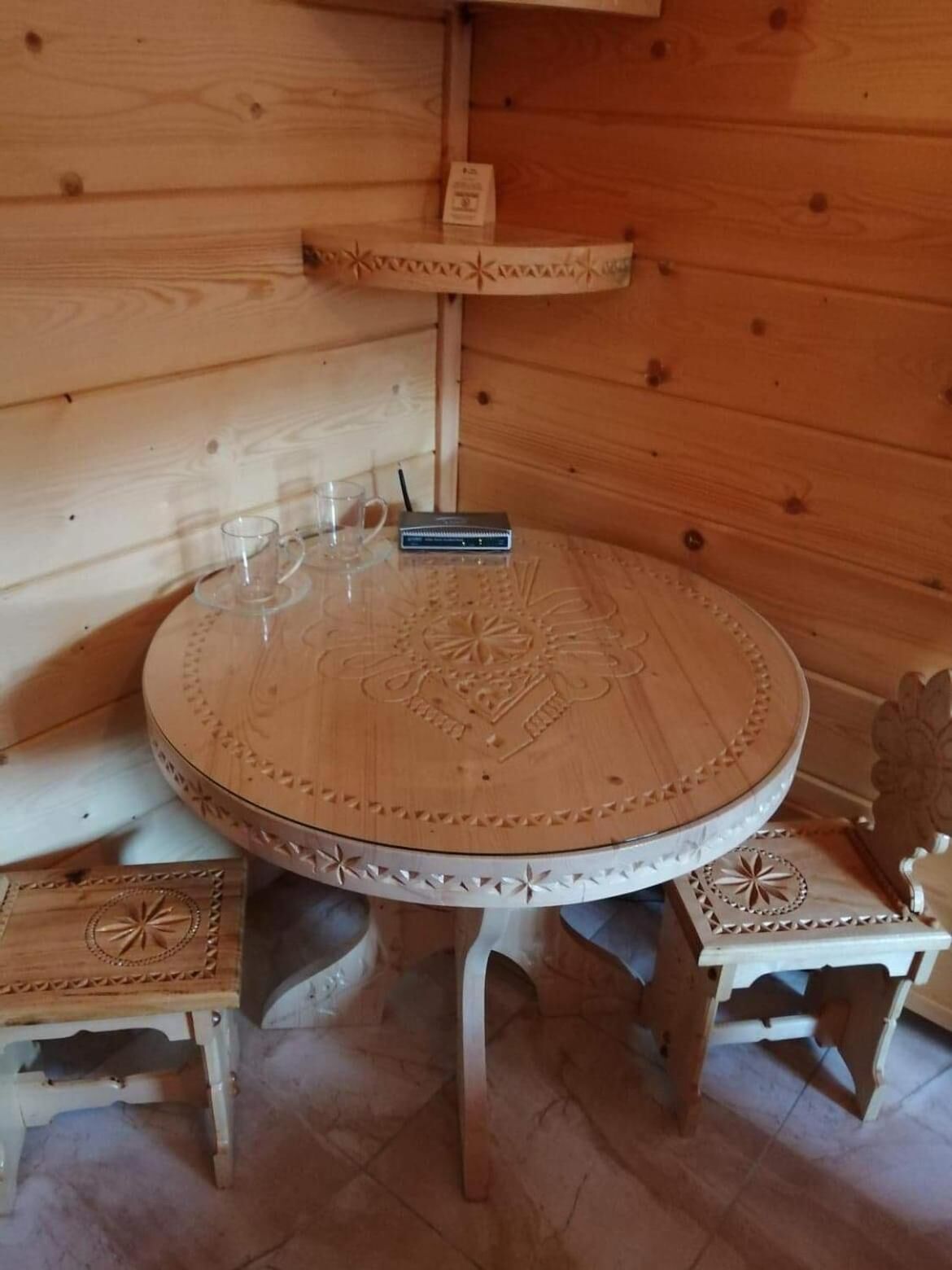 A wooden interior with a round table featuring intricate carvings, two matching chairs, and a small shelf with a book and a cup.