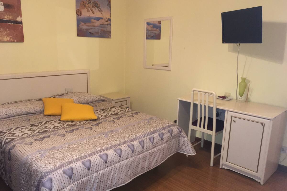 Un dormitorio con cama, escritorio, silla, espejo y televisor montado en la pared.