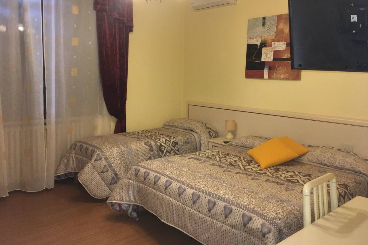 Habitación de hotel con dos camas, una ventana con cortinas, un equipo de aire acondicionado y una obra de arte colgada en la pared.