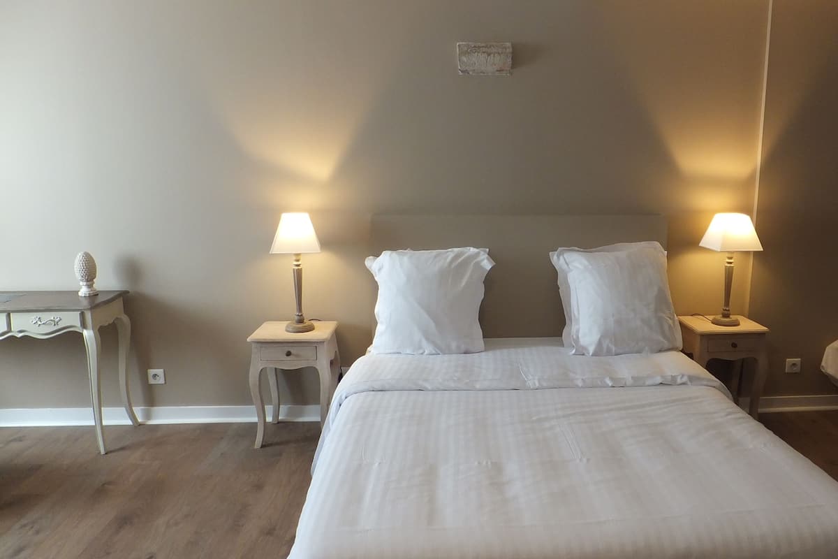 Een net opgemaakt bed met twee bedlampen, een klein nachtkastje met lamp, en een dressoir met decoratieve object.