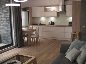 Une cuisine moderne avec des meubles blancs, un coin repas avec une table et des chaises en bois, et un canapé gris dans l’espace salon.