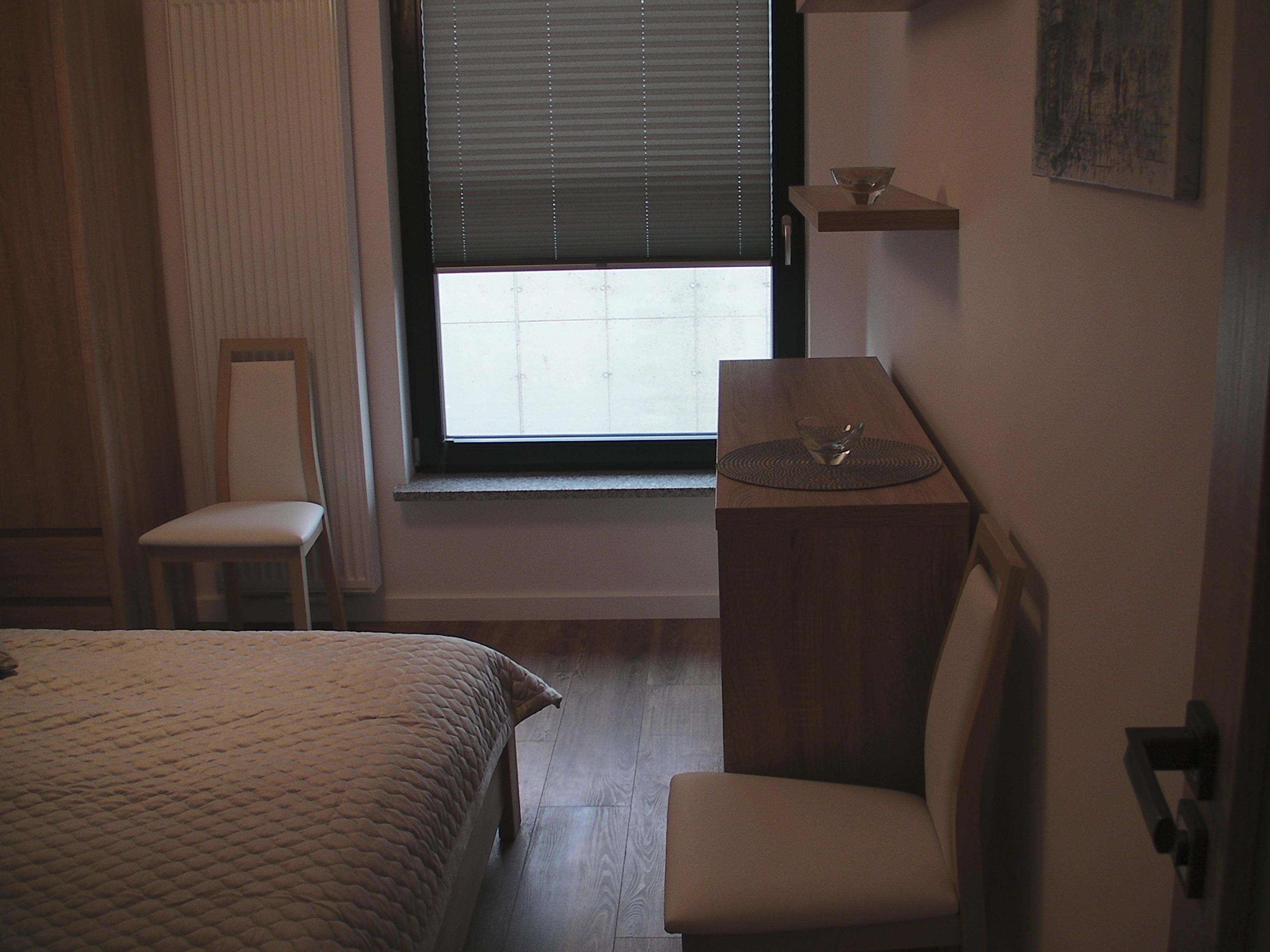 A bedroom with a bed, a chair, a window with blinds, a bedside table with a lamp, and a wall-mounted shelf.