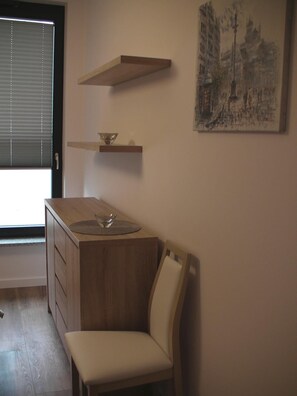Una habitación pequeña y acogedora con un escritorio de madera, una silla, una estantería con un cuenco y un cuadro en la pared.