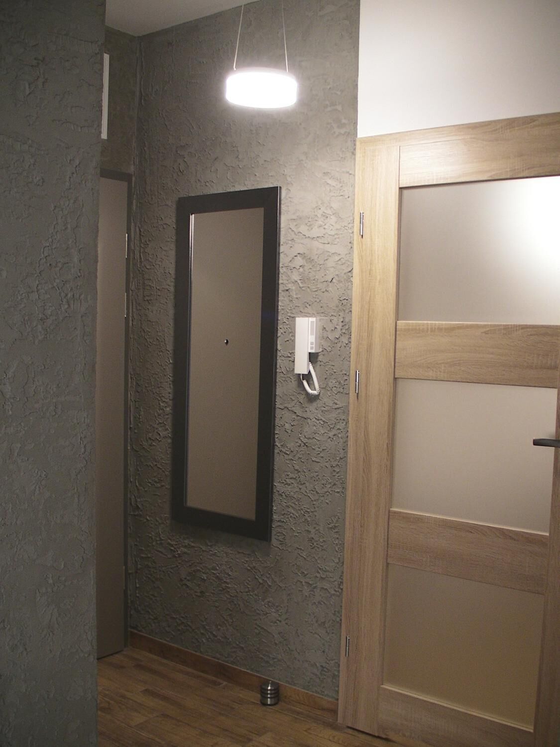 Una habitación con pared gris texturizada, una puerta de madera con paneles de vidrio esmerilado, una lámpara colgante y un pequeño recipiente metálico en el suelo.