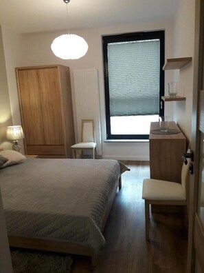 Un dormitorio con cama, una silla, armario, ventana con persianas y mesita de noche con lámpara.