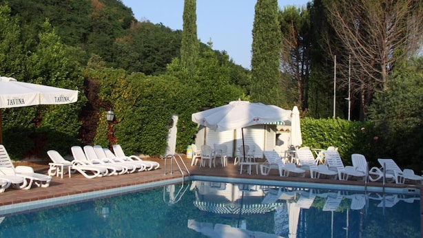 Una piscina con sillones de descanso y sombrillas, rodeada de árboles y una colina.