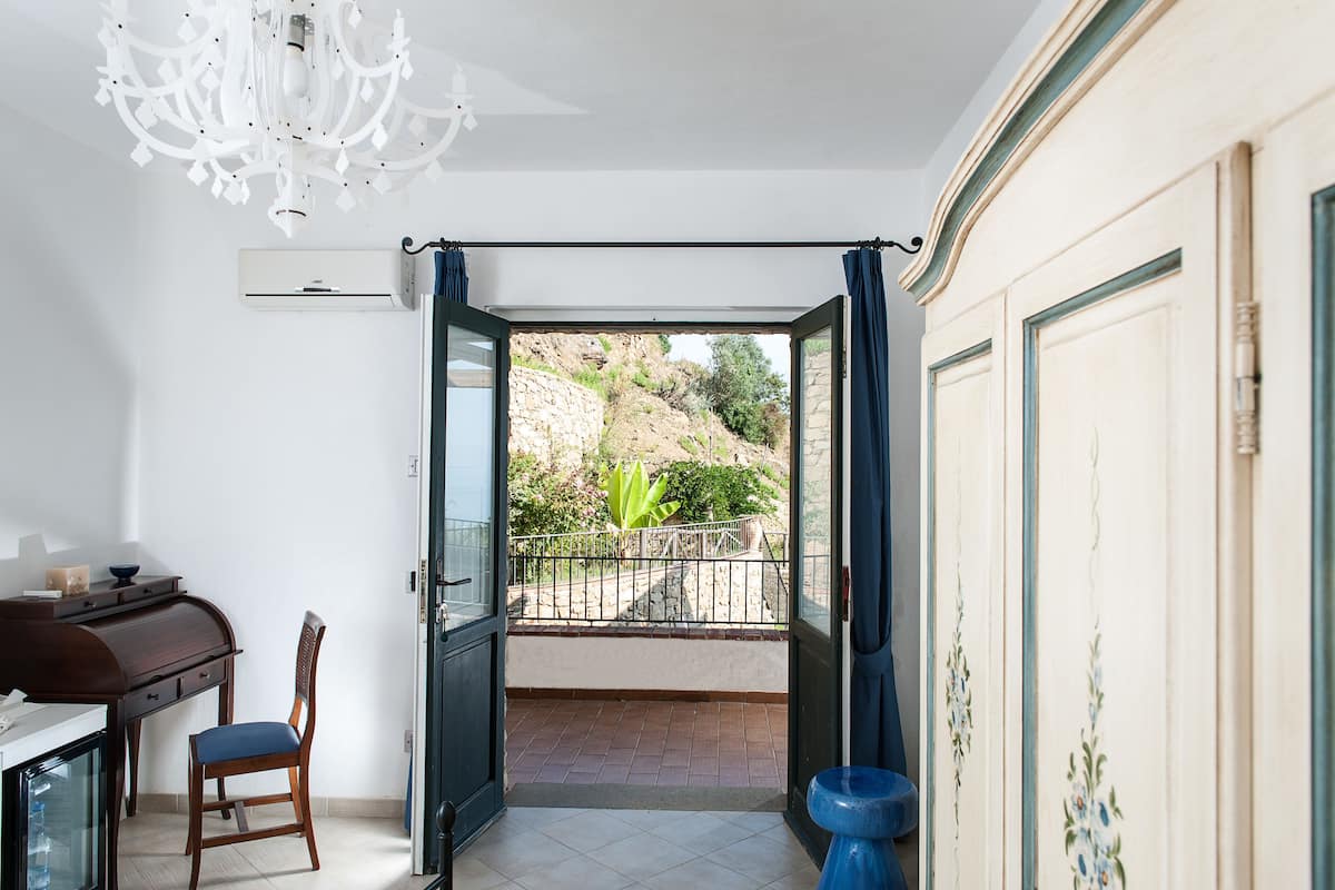 Una habitación con comedor, una silla, una mesa, una araña y una puerta abierta que da a un balcón con vistas a una ladera rocosa y vegetación.