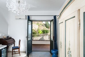 Ein Zimmer mit Essbereich, einem Sessel, einem Tisch, einer Kristallleuchter und einer offenen Tür, die auf einen Balkon mit Blick auf einen felsigen Hang und viel Grün führt.