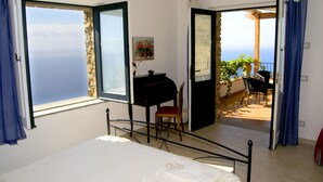 Quarto com cama, escrivaninha e cadeira. Há duas janelas com cortinas, uma das quais dá para uma varanda com vista para o mar.