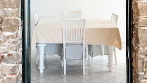 Uma área de refeições com parede de tijolos, mesa com toalha bege, cadeiras brancas e luminária pendente.
