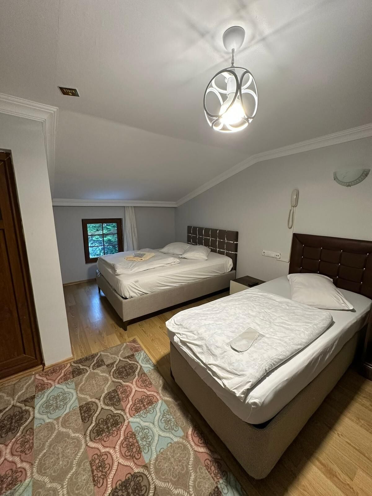 A bedroom with two beds, a patterned rug, a ceiling light fixture, and a window with curtains.