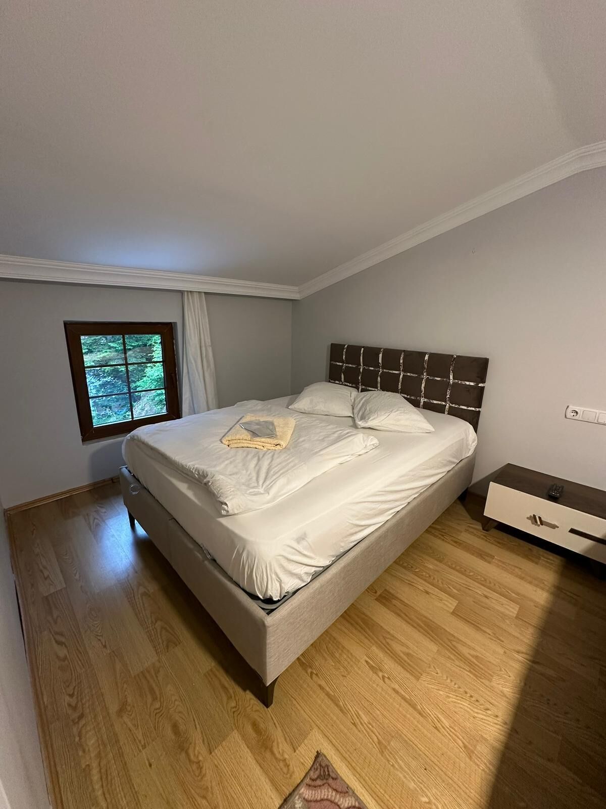 A bedroom with a bed, a window, a nightstand, and a wooden floor.