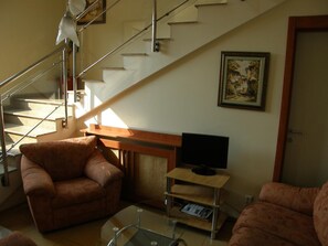 Een woonkamer met een bank, een televisie op een houten dressoir, een trap en een schilderij aan de muur.
