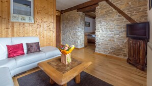 A cozy living room with a wooden sofa, a coffee table with a bouquet, a stone wall, and a television. - La ferme Morin la Ronde Fontaine (Fournet-Blancheroche)