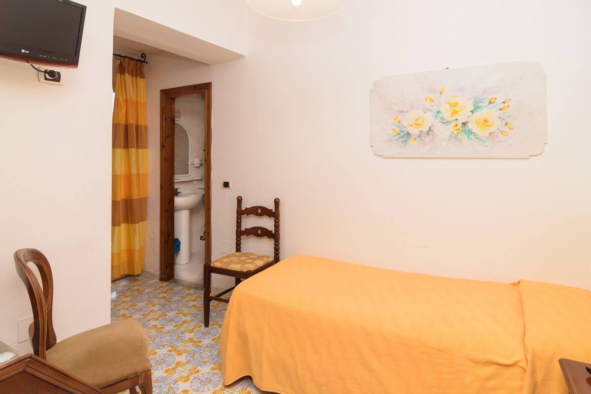 Een kleine kamer met een bed, een stoel, een schilderij en een televisie.