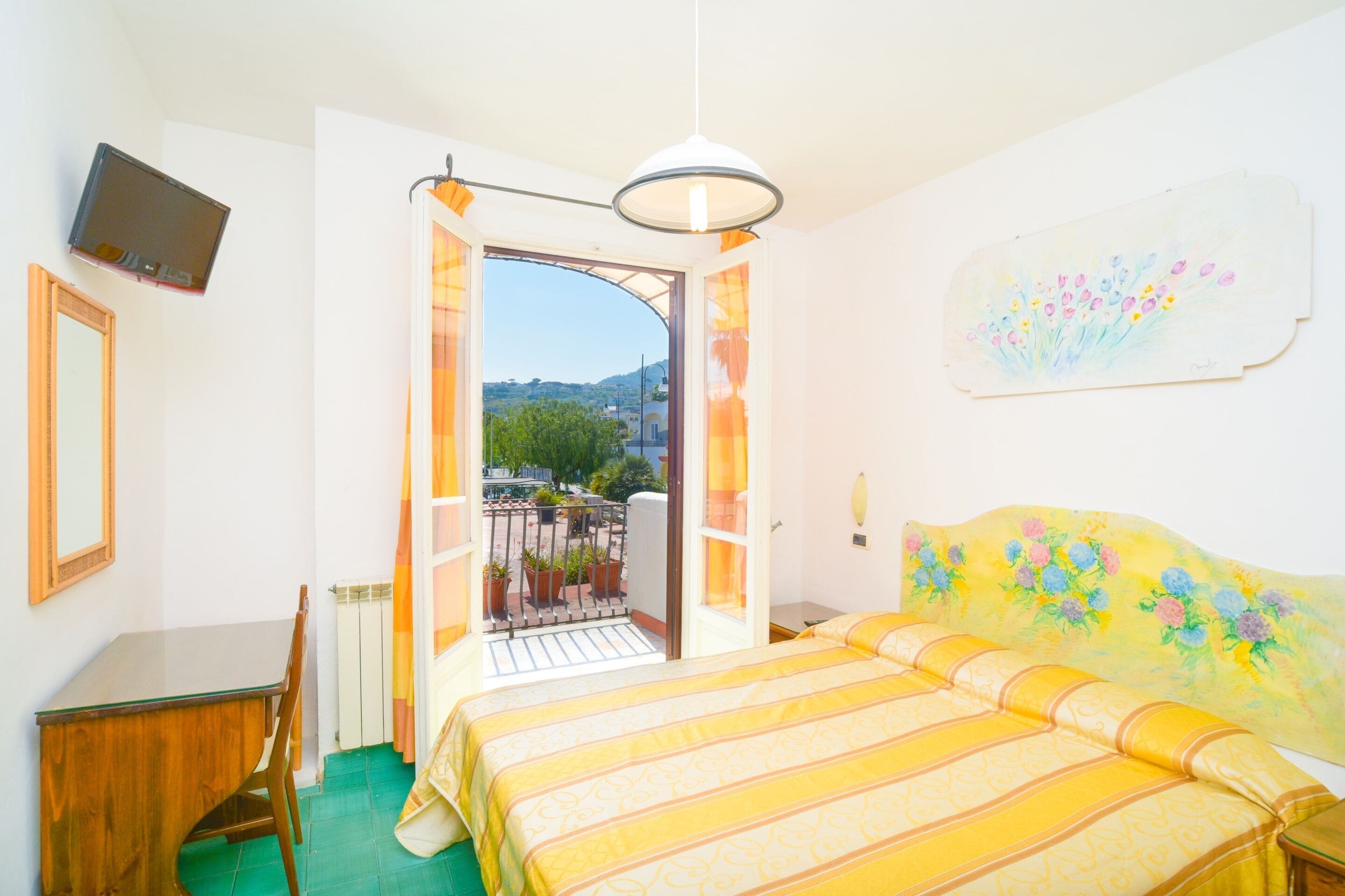 A bedroom with a bed, a desk, a chair, a TV, and a balcony with a view of the city.