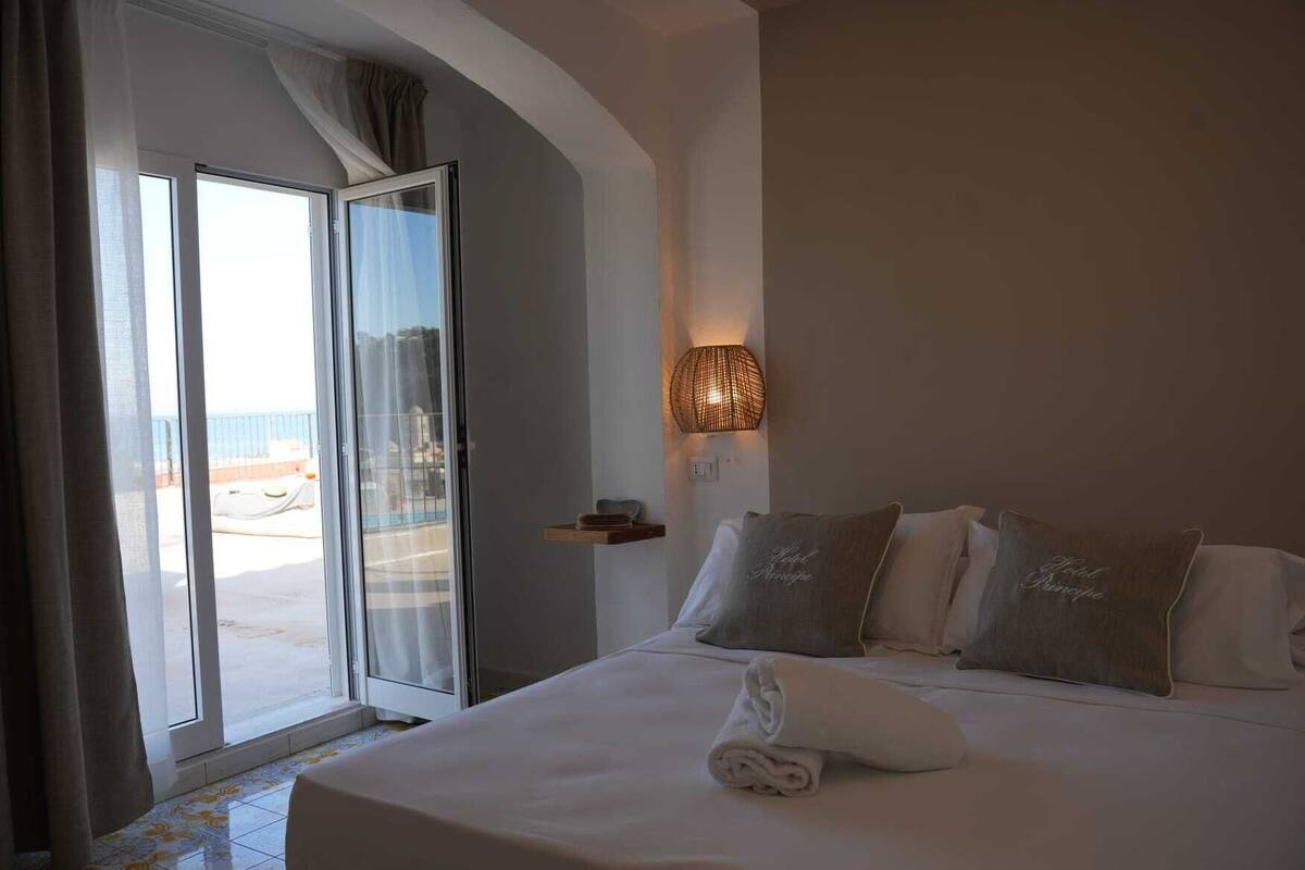 Een hotelkamer met een bed, nachtkastje, lamp en uitzicht op het strand door een schuifdeur.