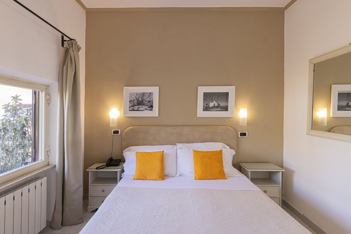 A hotel room with a large bed, two bedside tables, a nightstand, a window with curtains, and three wall-mounted picture frames.
