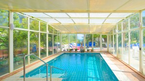 A covered indoor swimming pool with clear glass walls, a tiled floor, and a view of the outside.